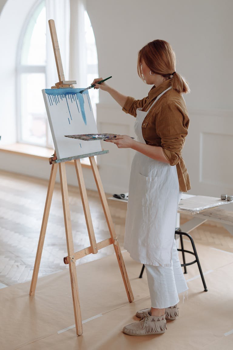 Female artist painting on canvas in sunny studio with easel and brush.